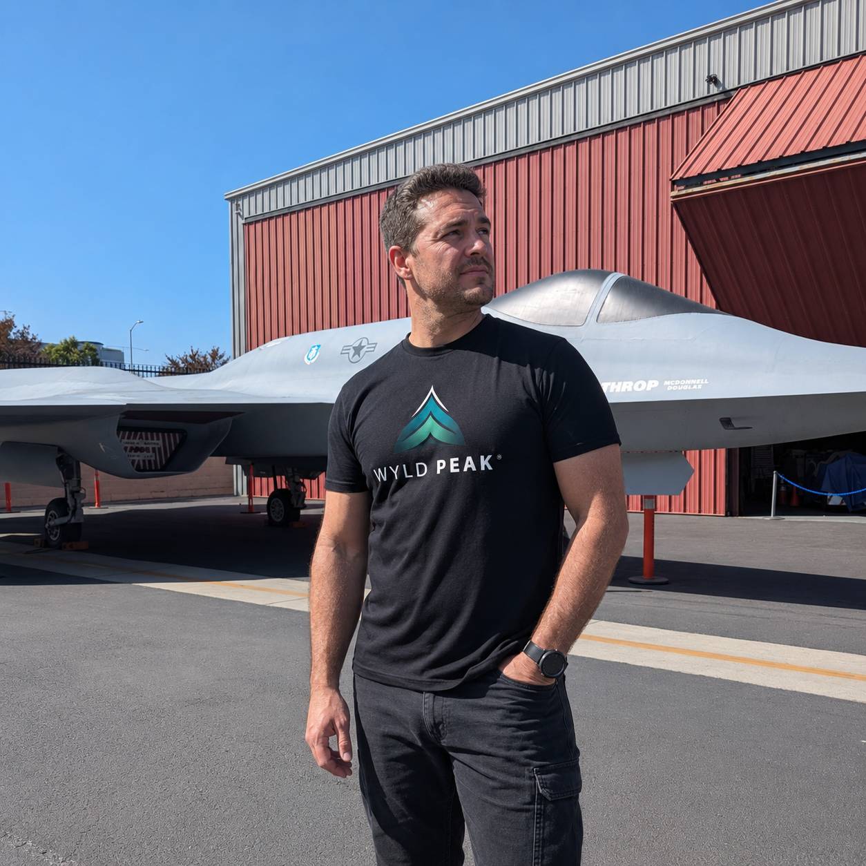 A man wearing a black t-shirt with a green and white 'WYLD PEAK' logo stands in front of a gray yf-23 black widow military jet and a red hangar under a clear blue sky in LA.