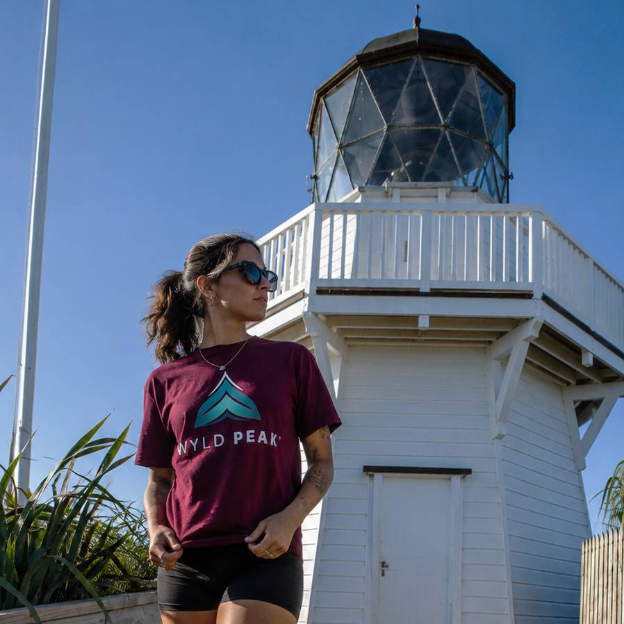 A woman wearing a maroon t-shirt with a teal and white 'WYLD PEAK' mountain logo stands in front of a white lighthouse under a clear blue sky.