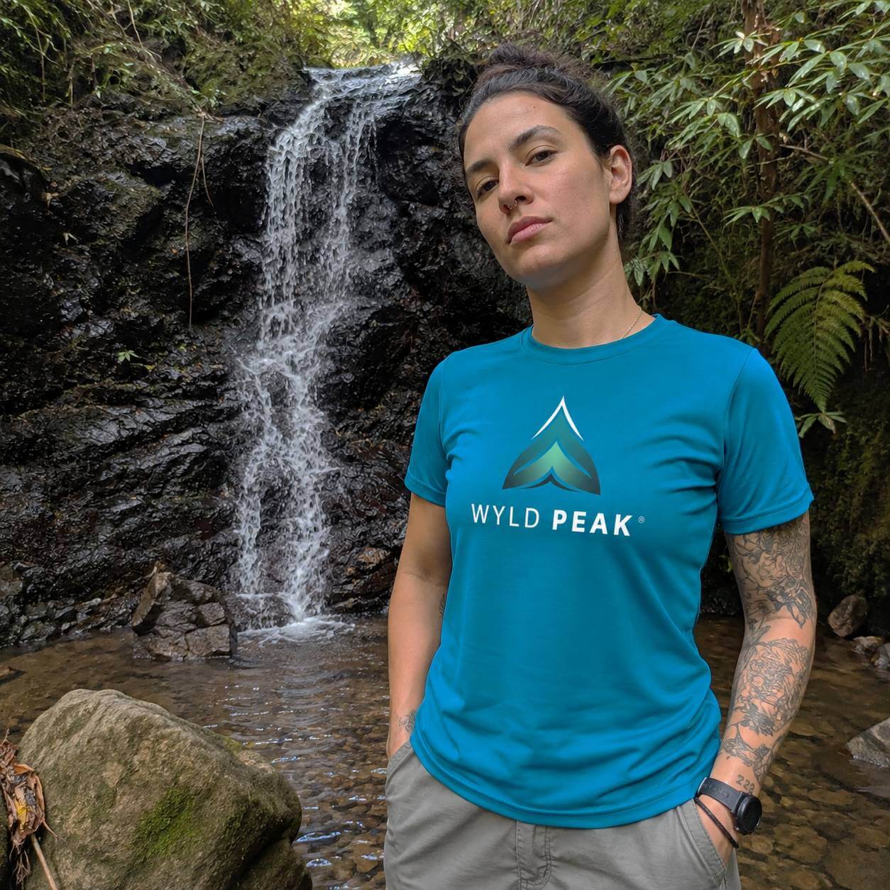 A woman standing outdoors in front of a small waterfall and forest foliage, wearing a bright teal T-shirt with a mountain logo and the text 'WYLD PEAK' on the chest, paired with gray shorts.