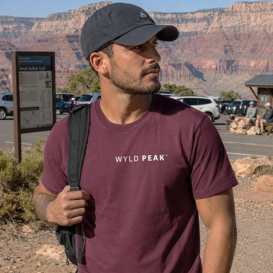 Man wearing a maroon 'WYLD PEAK' t-shirt with a backpack in a desert setting.