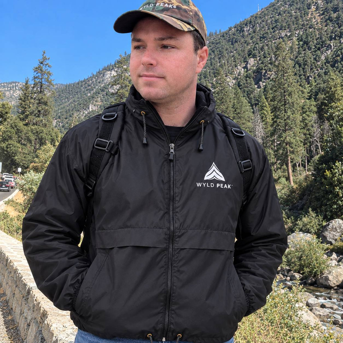 A man wearing a black waterproof jacket with a white 'WYLD PEAK' logo on the chest, standing outdoors in front of a mountain backdrop.