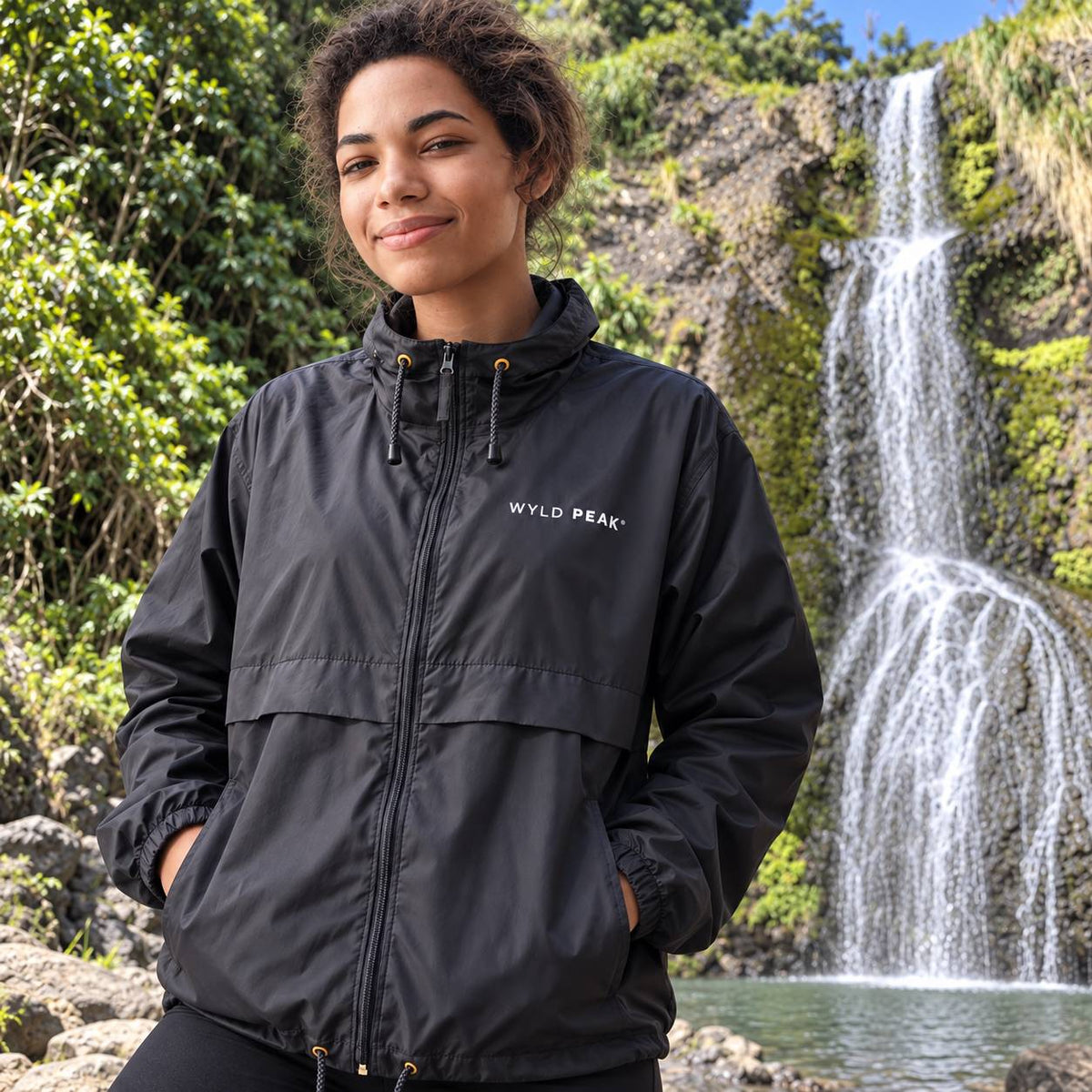 A woman wearing a black waterproof jacket with 'WYLD PEAK.' printed in white on the chest, standing outdoors in front of a waterfall and lush green forest.