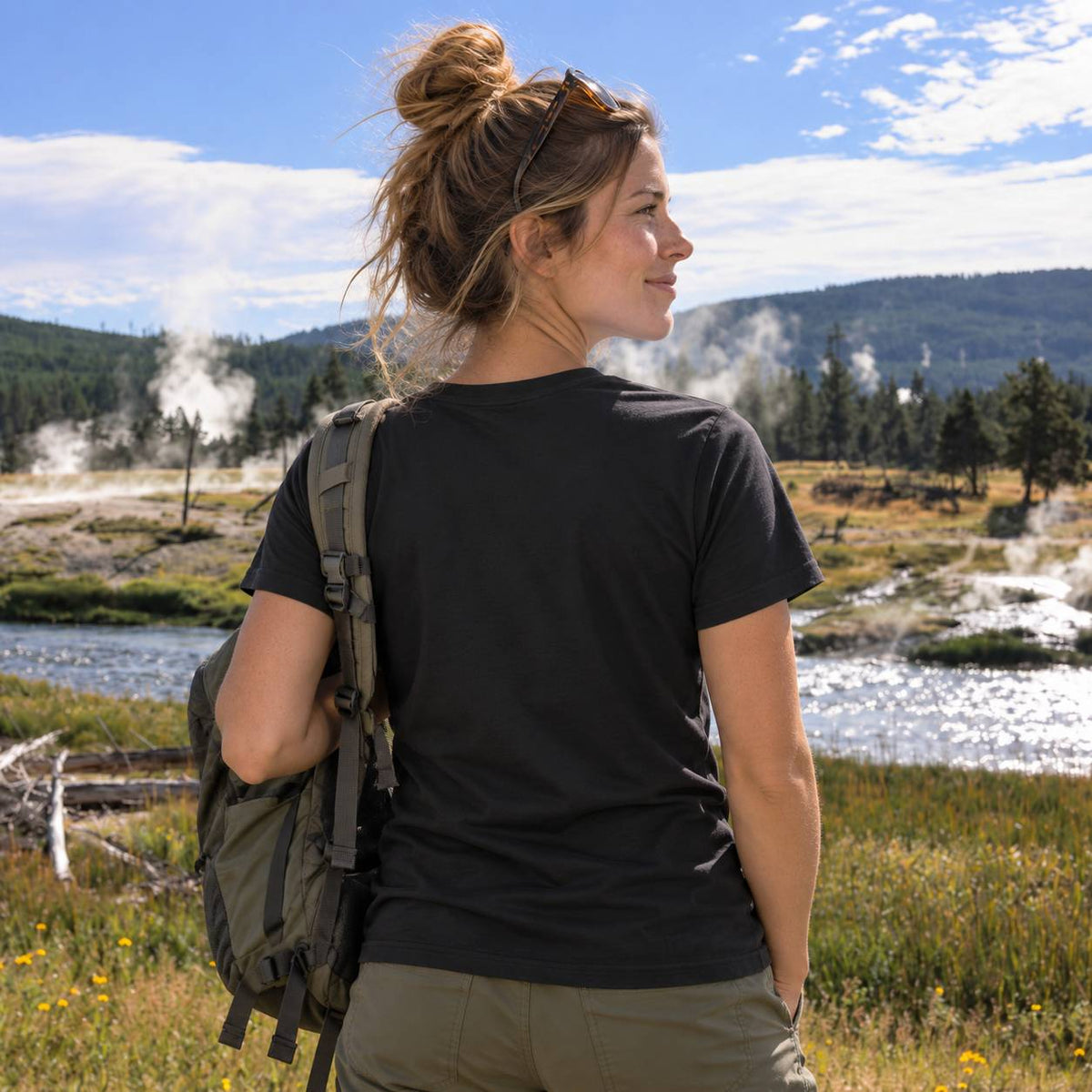 A woman hiking outdoors wearing a black short-sleeve t-shirt, olive green pants, and carrying an olive green backpack, photographed from behind with a scenic landscape of a river and mountains in the background.