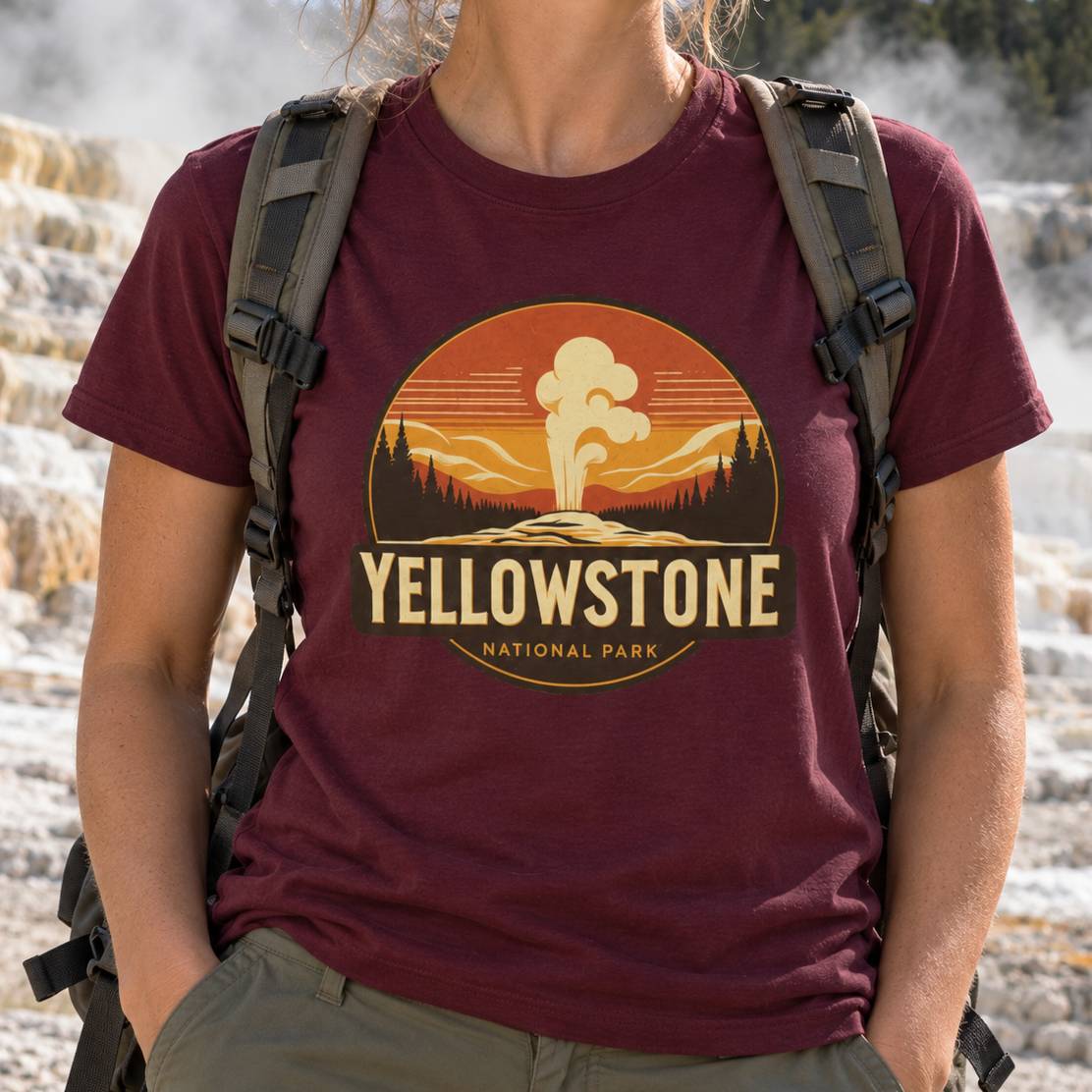 A person wearing a maroon t-shirt with a retro-style Yellowstone National Park graphic, carrying a hiking backpack in an outdoor geothermal setting.