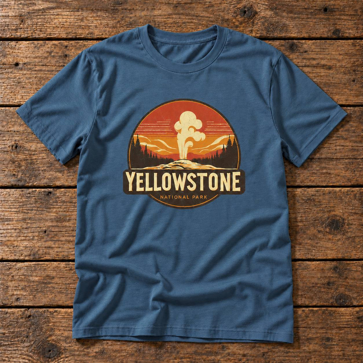 A blue crewneck t-shirt featuring a circular Yellowstone National Park graphic on the chest, laid flat on a rustic wooden plank background.