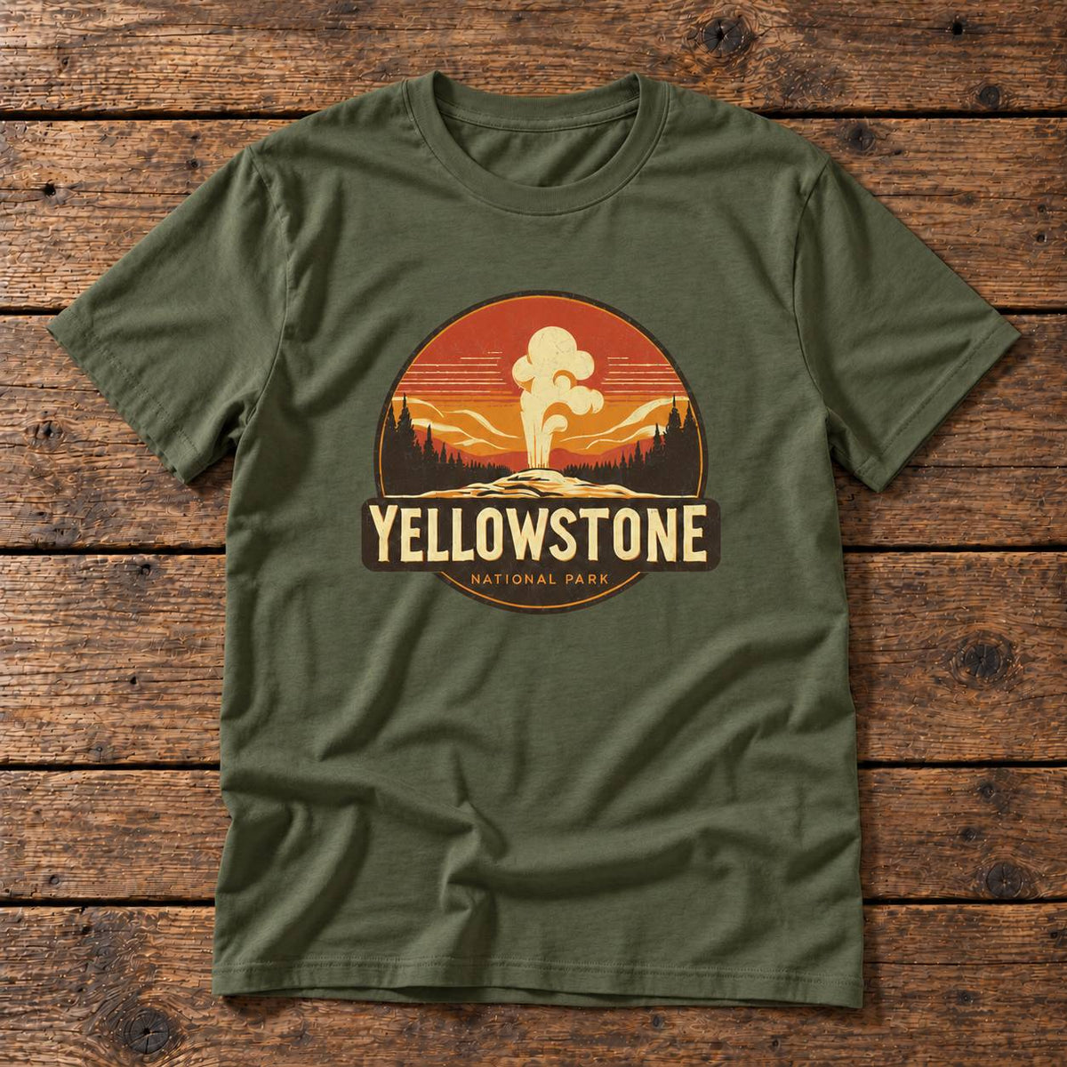 An olive green crewneck t-shirt featuring a retro circular Yellowstone National Park graphic on the chest, laid flat on a rustic wooden plank background.