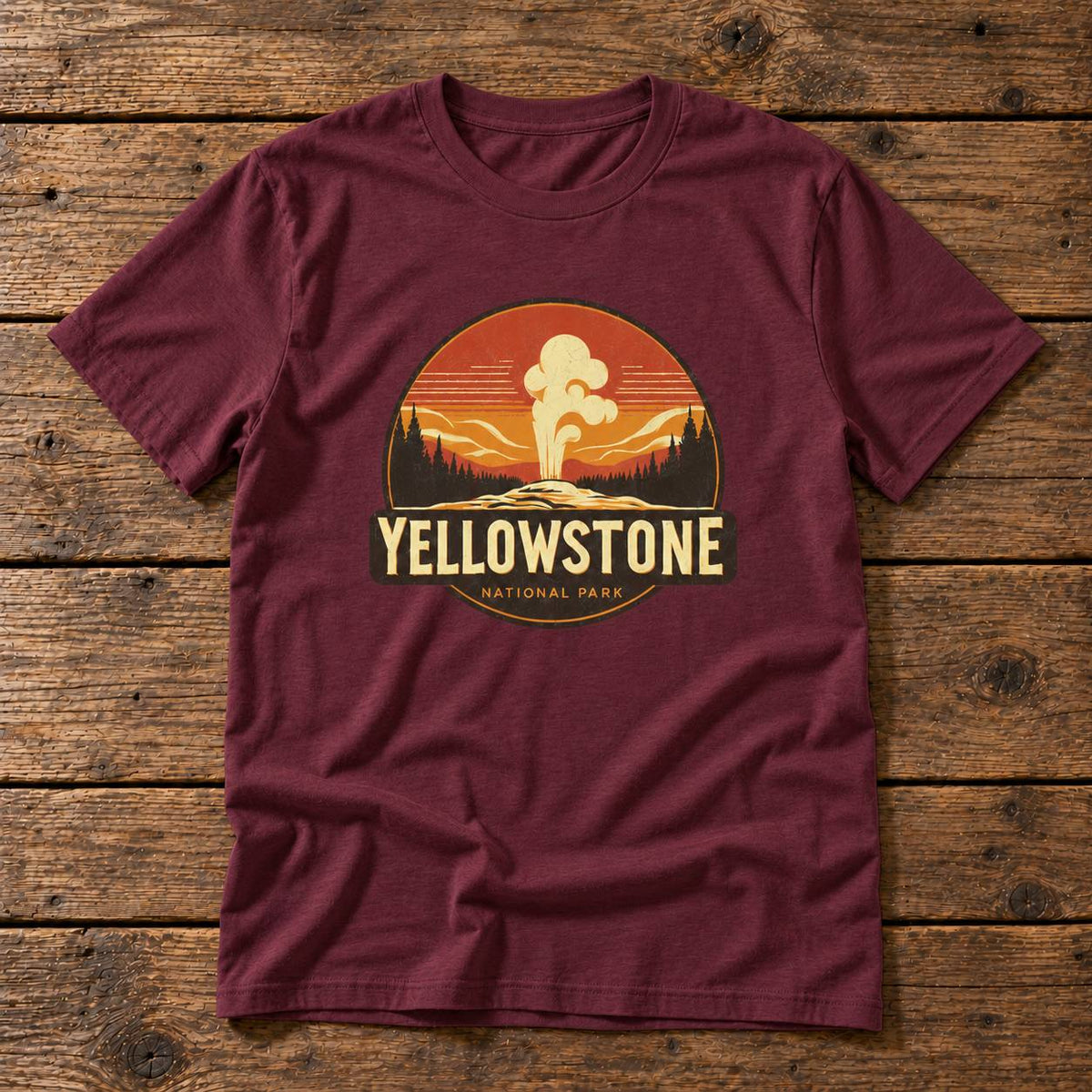A maroon short-sleeve crewneck t-shirt laid flat on a rustic wooden plank background. The center of the shirt features a circular graphic print depicting a Yellowstone National Park sunset scene with a hot spring, and the words 'YELLOWSTONE NATIONAL PARK' printed below it.