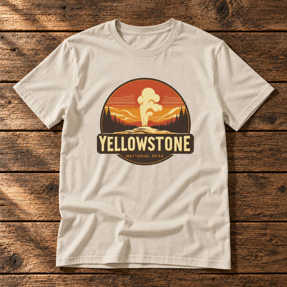 A light beige crewneck t-shirt laid flat on a rustic dark wood background. The shirt features a circular graphic print in the center depicting a Yellowstone National Park sunset scene with a hot spring, and the words 'YELLOWSTONE NATIONAL PARK' printed below it.