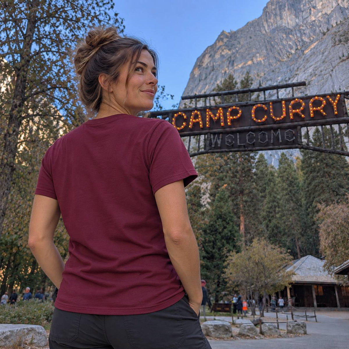 A woman wearing a maroon short-sleeve t-shirt and dark gray pants stands in front of a 'Camp Curry Welcome' sign at Yosemite National Park, looking back over her shoulder with a mountain in the background.