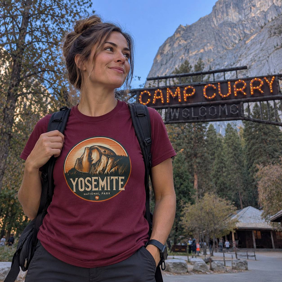 A woman wearing a maroon t-shirt with a Yosemite National Park graphic stands in front of the 'Camp Curry Welcome' sign in Yosemite Valley, holding a black backpack.