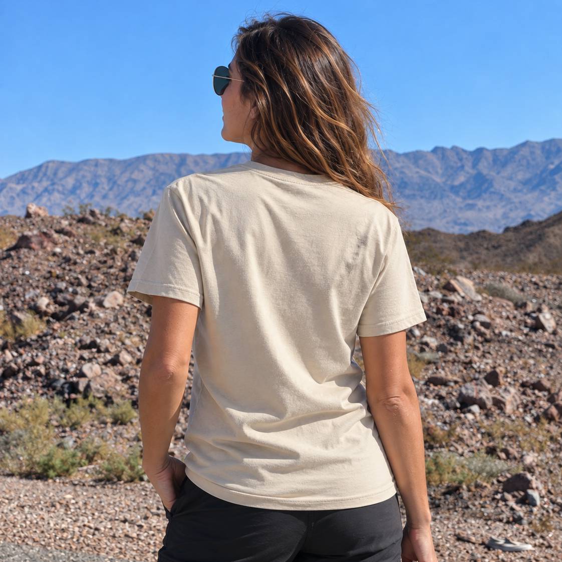 A woman wearing a light beige short-sleeve t-shirt and black pants, photographed from behind in an outdoor desert landscape with mountains and a blue sky.