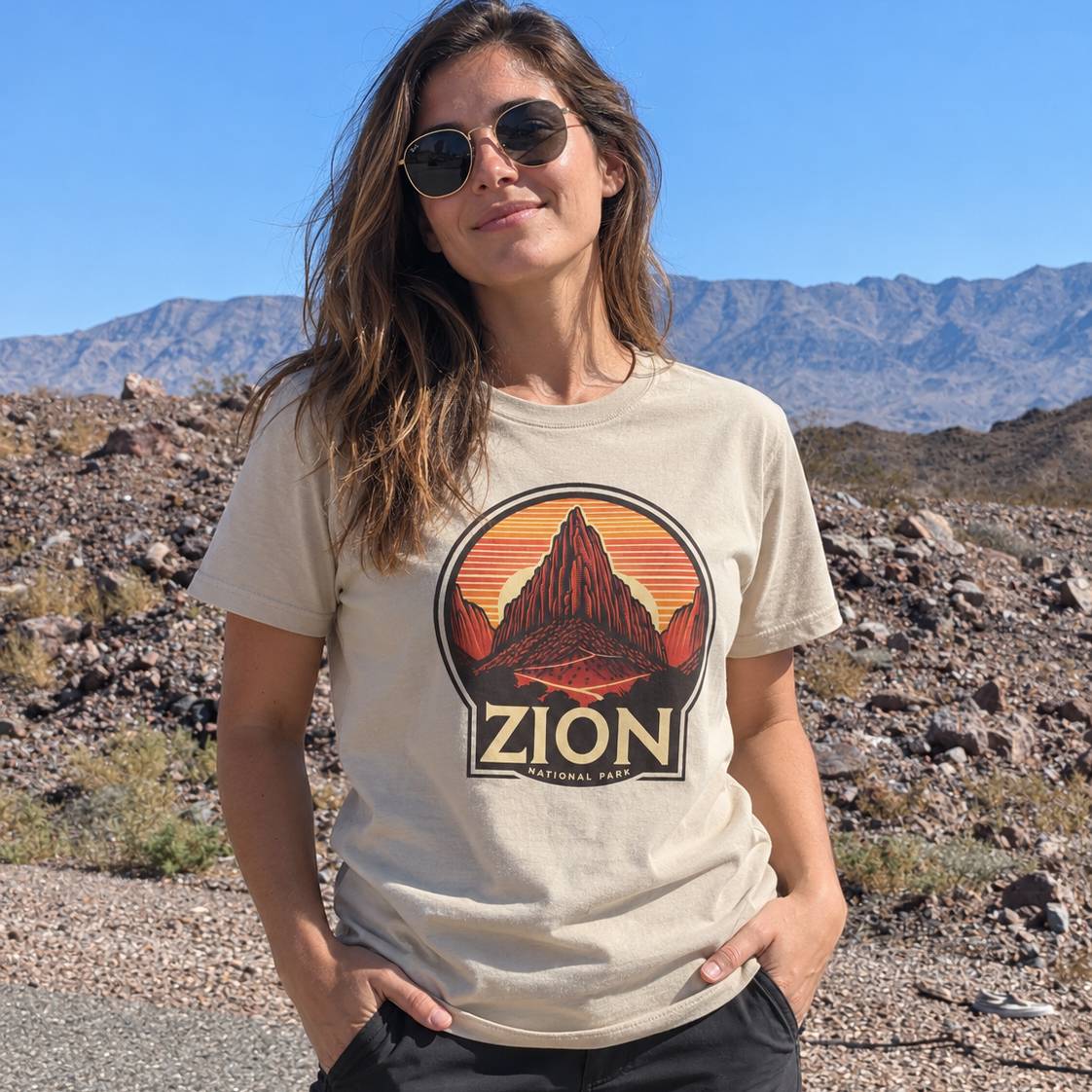 A woman wearing a beige graphic t-shirt with a 'Zion National Park' sunset design, standing outdoors in a rocky desert landscape with mountains in the background.