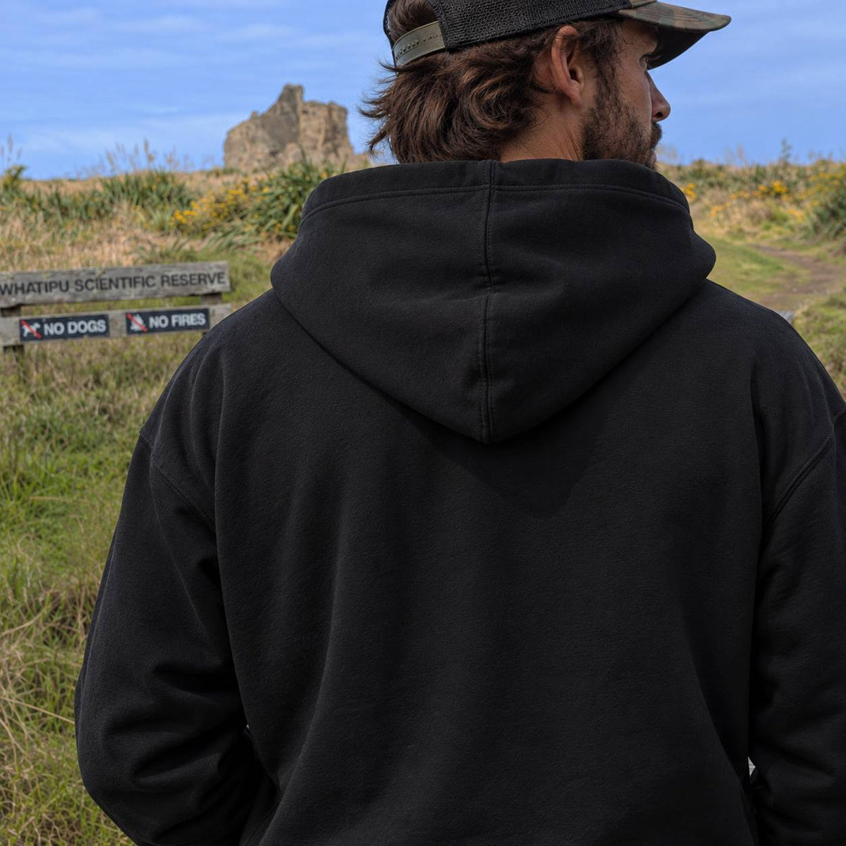 A man viewed from behind wearing a black pullover hoodie and a dark baseball cap, standing outdoors in a grassy field with a sign reading 'WHATIPU SCIENTIFIC RESERVE' and 'NO DOGS NO FIRES' to the left.