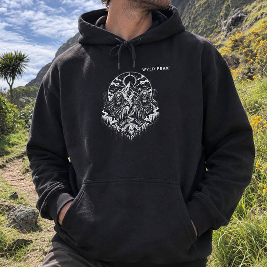 A man wearing a black pullover hoodie with a white graphic print of three figures in front of a mountain peak, set in an outdoor hiker's environment.