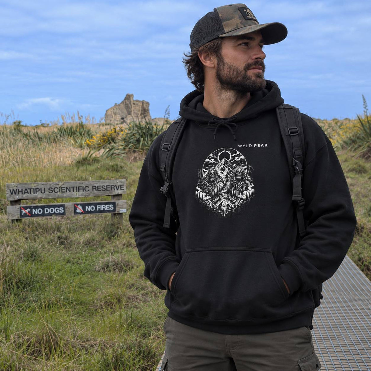 A man wearing a black pullover hoodie with a white graphic print of a wolf and the text 'WYLD PEAK' stands outdoors in a grassy field under a blue sky. He is also wearing a camouflage baseball cap and a black backpack.
