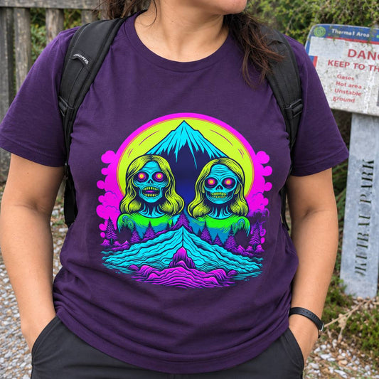 A person wearing a purple short-sleeve graphic t-shirt with a vibrant neon-style print of two smiling skull-faced women with long blonde hair in front of a mountain. The person is also wearing a black backpack and black pants, standing outdoors near a trail sign.