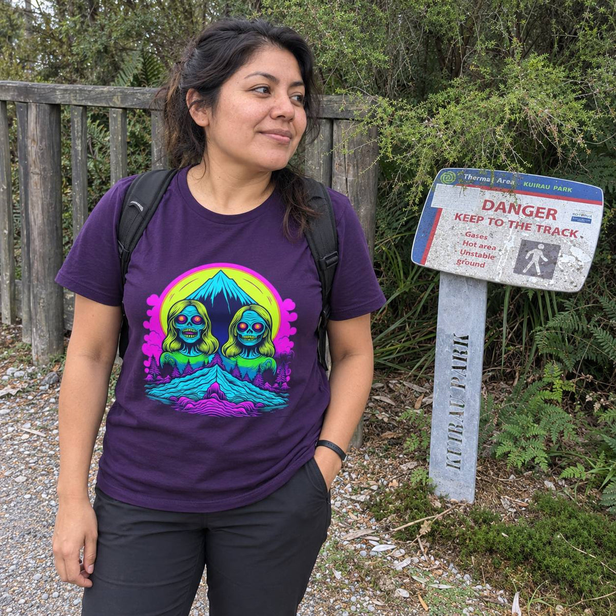A woman wearing a purple t-shirt with a colorful graphic print stands outdoors next to a trail sign. The graphic features two cartoonish green creatures inside a neon yellow and pink circular design.