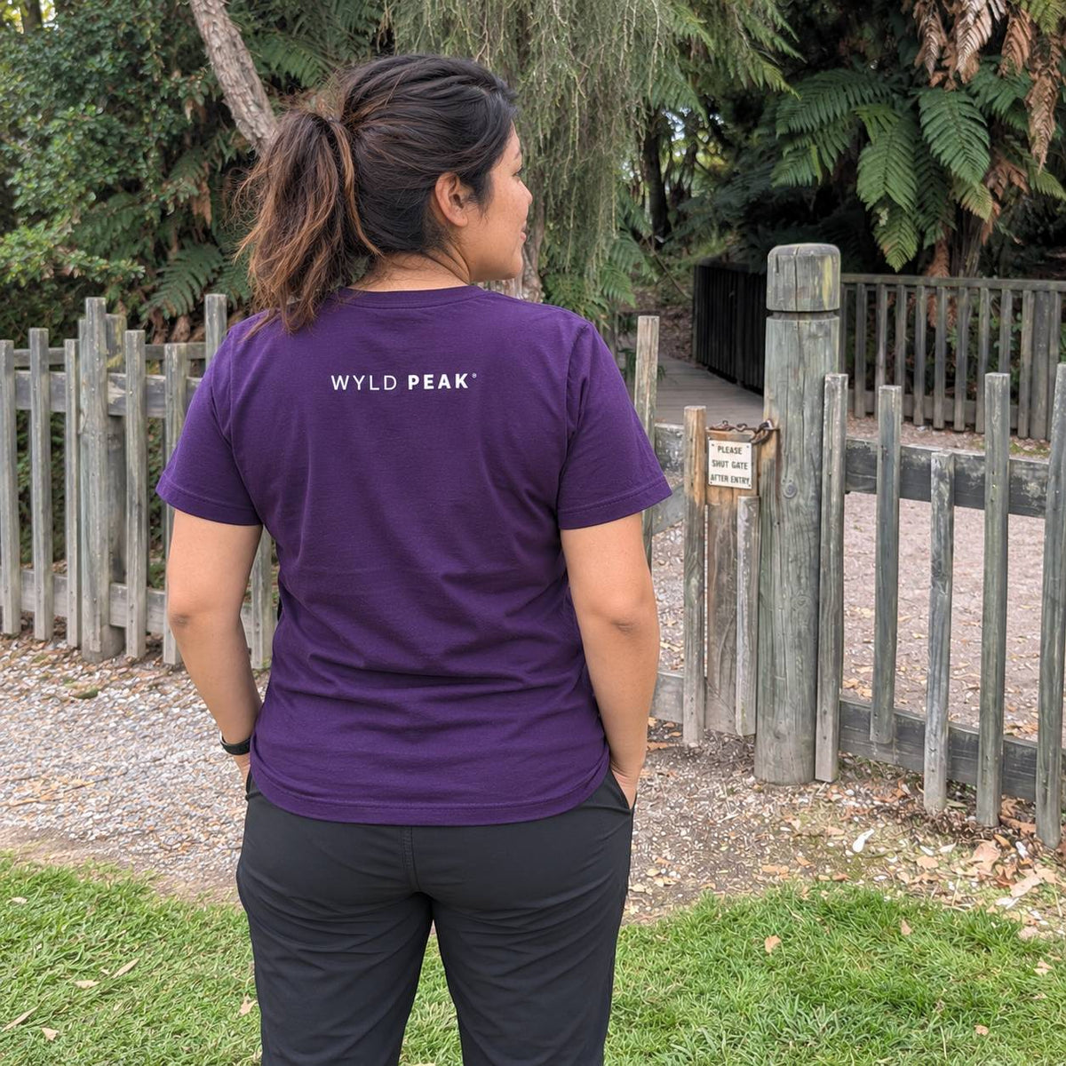 A woman viewed from behind wearing a purple t-shirt with 'WYLD PEAK' printed in white on the back, standing outdoors in front of a wooden fence and trees.