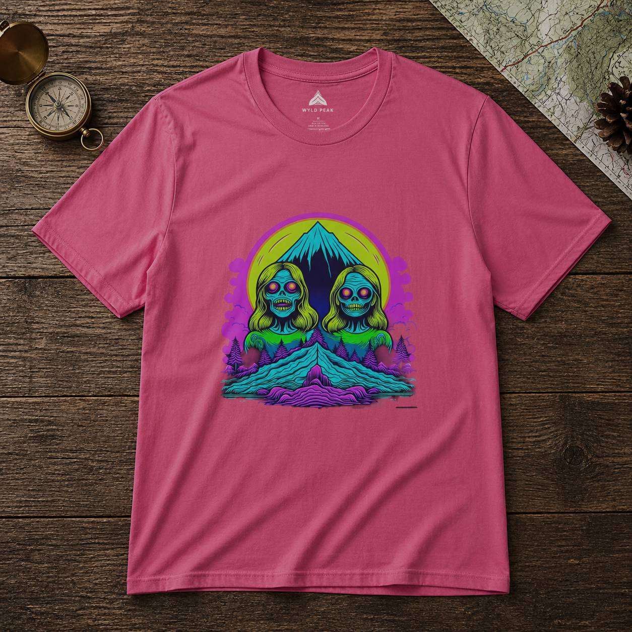 Pink t-shirt with a graphic design of two green characters in front of a mountain on a wooden surface.
