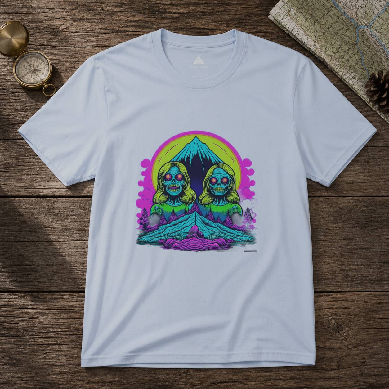 White t-shirt with a colorful graphic design of two green characters and a mountain on a wooden surface.
