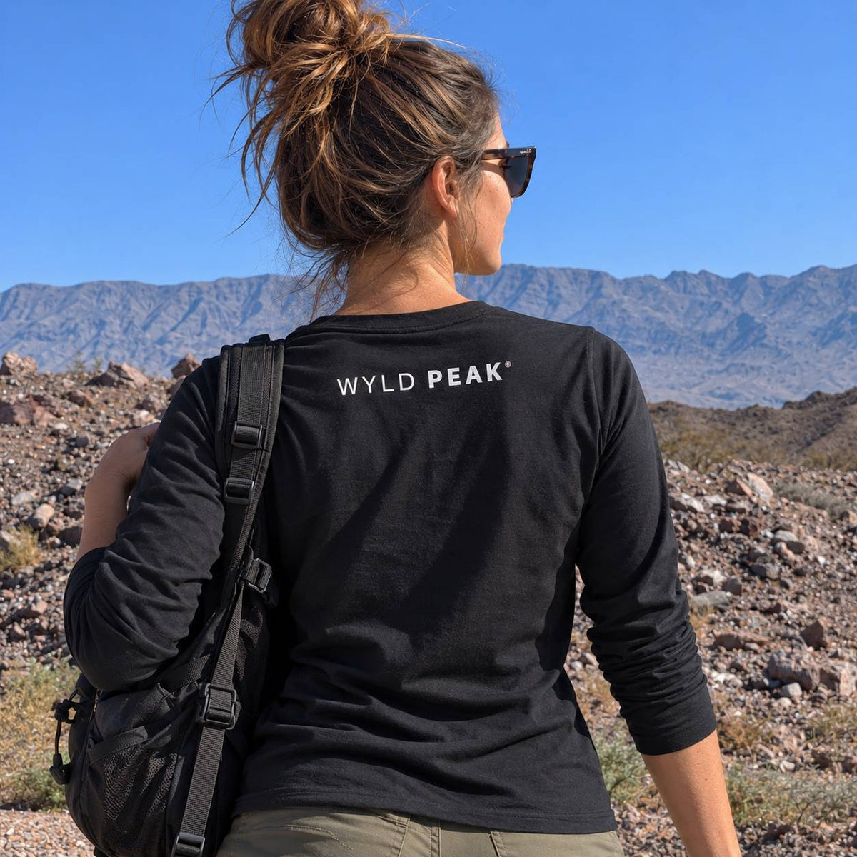 A woman seen from behind wearing a black long-sleeve t-shirt with 'WYLD PEAK' printed in white on the back, set against a desert landscape with mountains and a blue sky.
