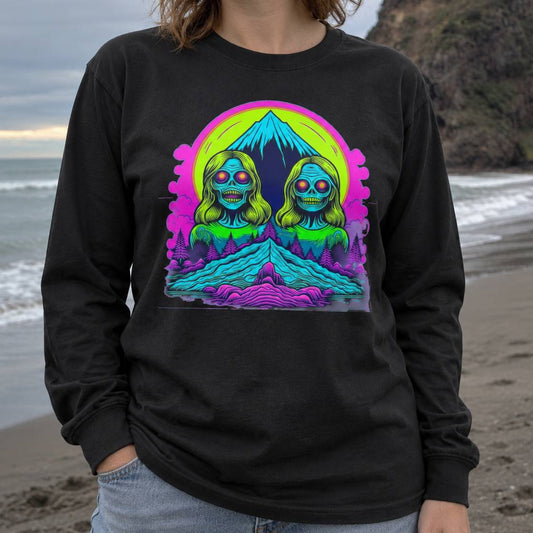 A person standing on a beach wearing a black long-sleeve t-shirt with a colorful retro graphic of two green and blue alien-like figures in front of a neon pink and yellow sun and mountain.