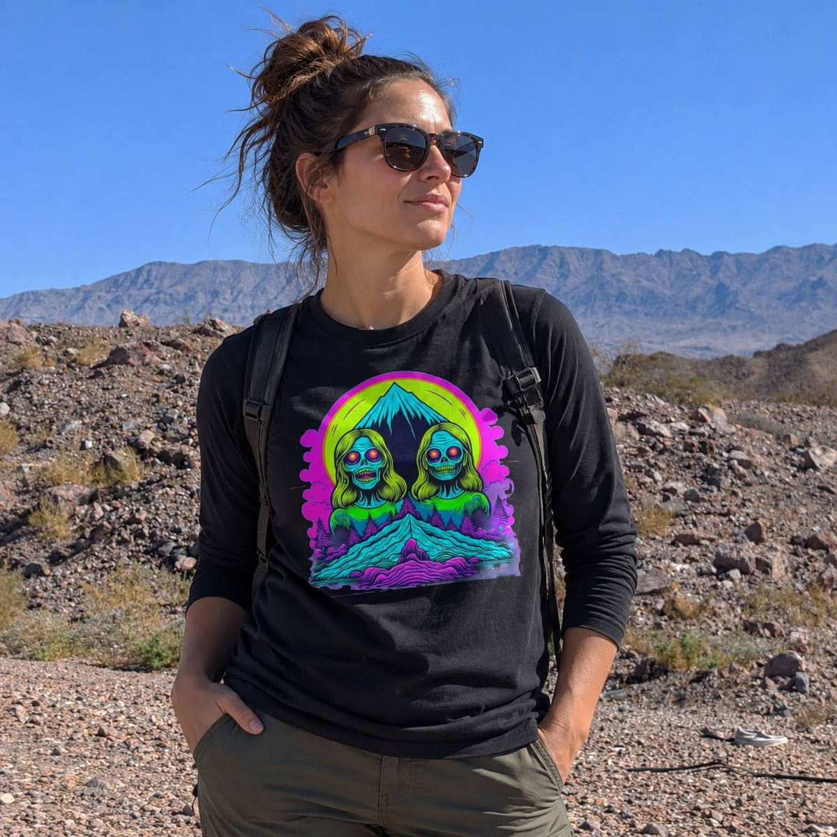 A woman wearing a black long-sleeve t-shirt with a colorful psychedelic graphic of two green aliens under a pink and yellow moon, standing in a rocky desert landscape with mountains in the background.