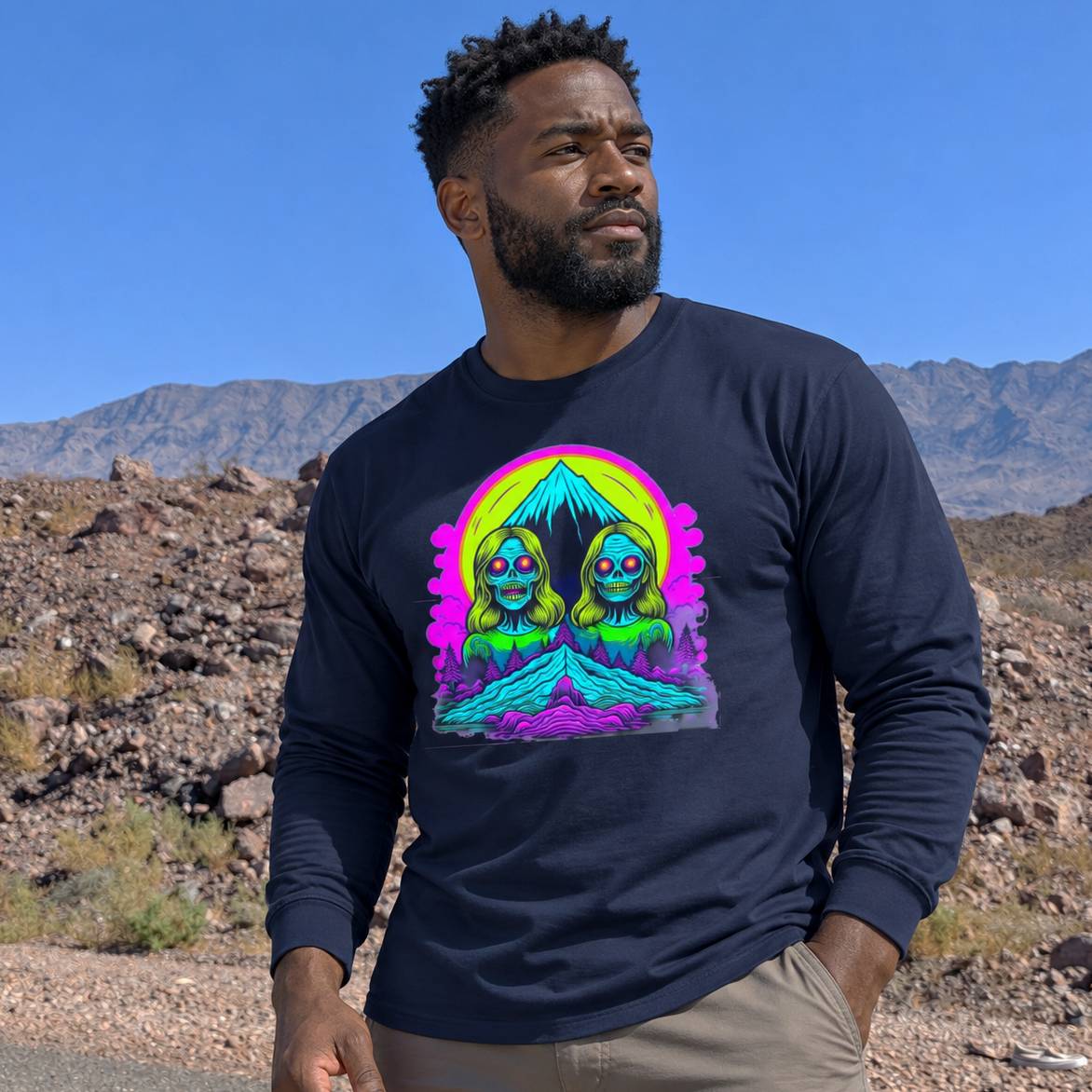 A man wearing a navy blue long-sleeve t-shirt with a vibrant, colorful graphic print of two green aliens under a pink and yellow moon, standing outdoors in a desert landscape with mountains in the background.