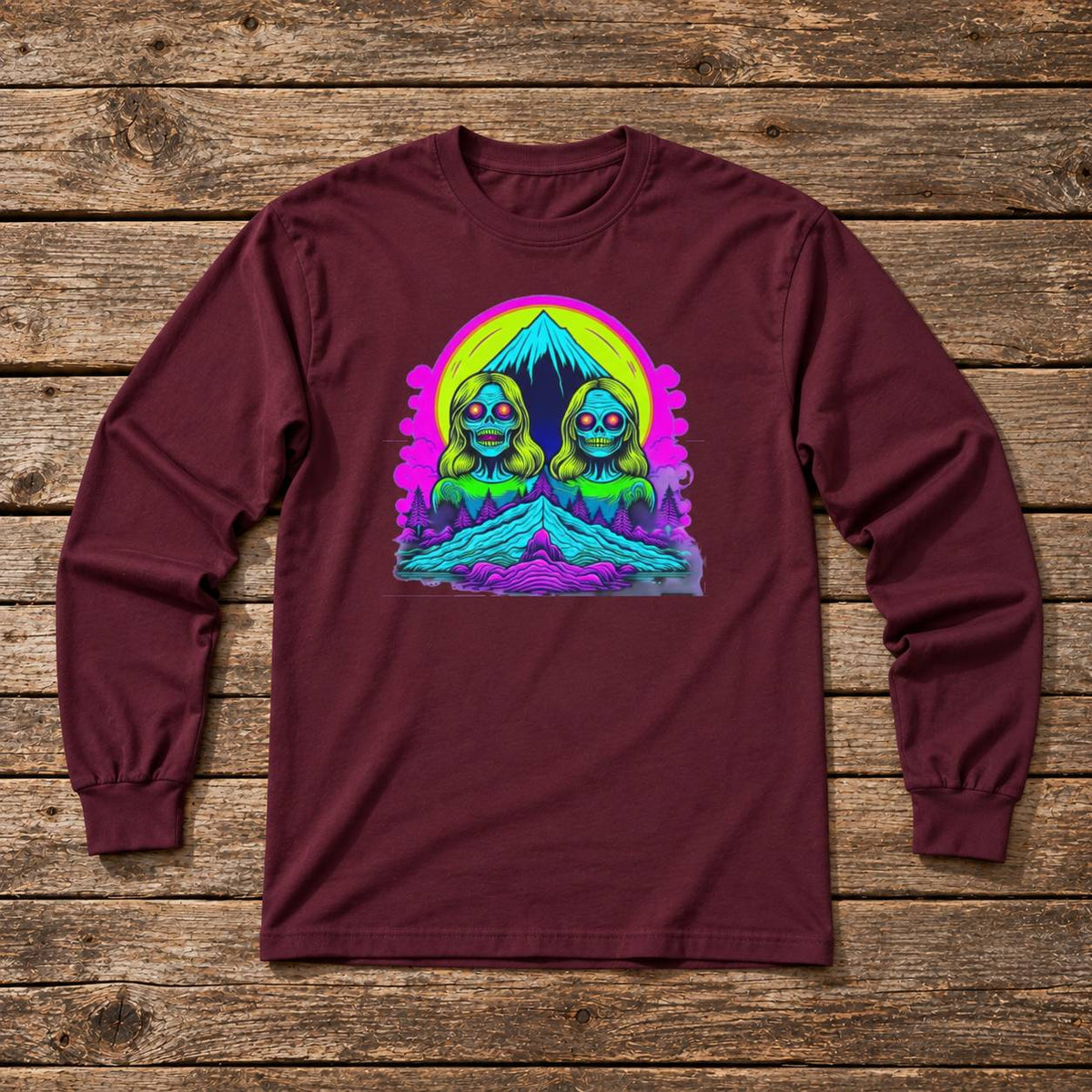 A long-sleeve crewneck t-shirt in maroon, laid flat on a rustic wooden plank background. The shirt features a colorful retro graphic on the chest of two green alien-like figures with pink sunglasses in front of a neon yellow and purple mountain background.