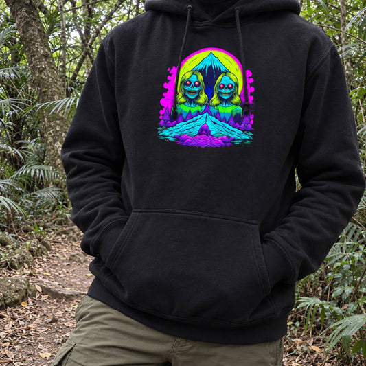 A person wearing a black pullover hoodie with a colorful psychedelic graphic of two mirrored cartoon figures in a neon landscape, photographed outdoors in a forest setting.