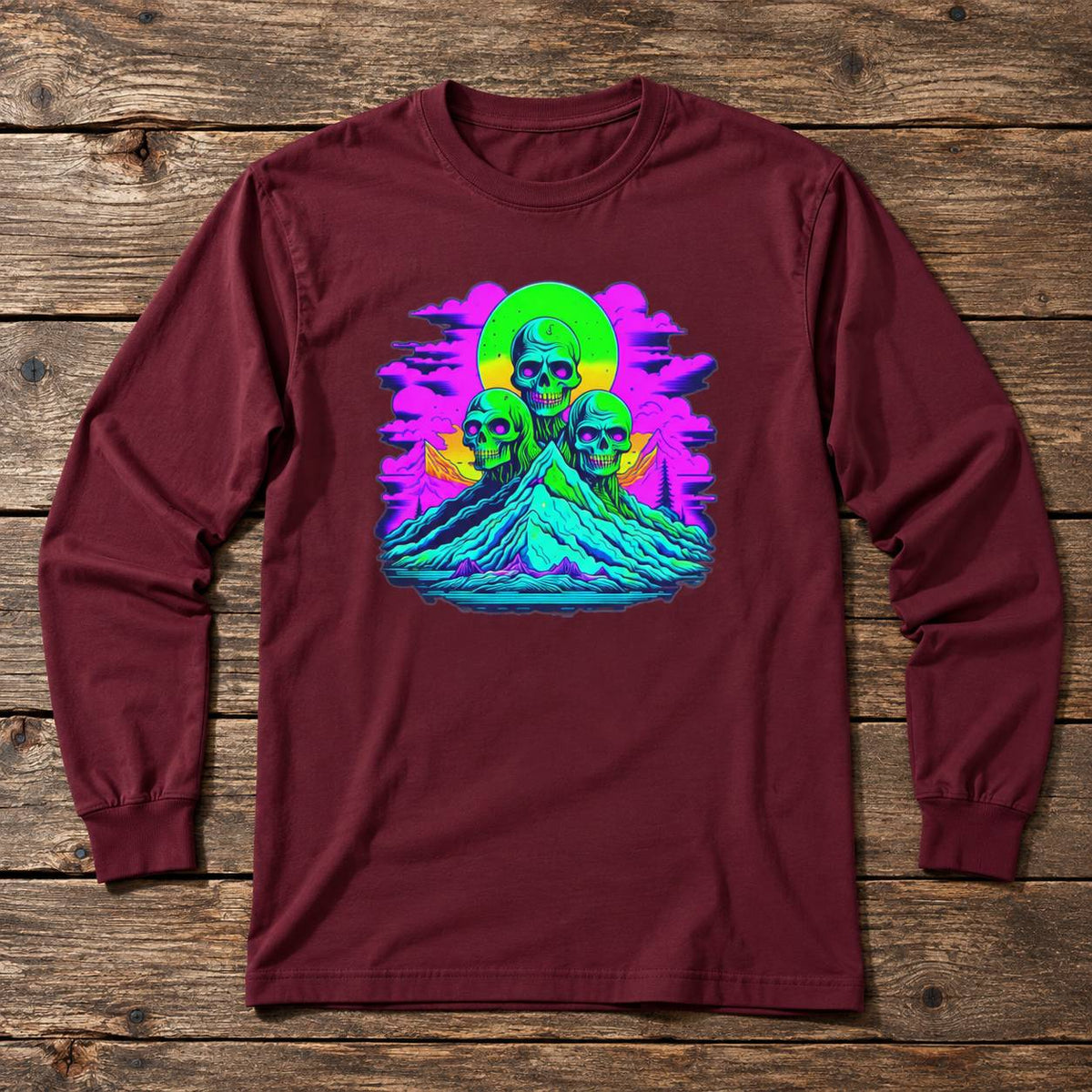 A long-sleeve crewneck t-shirt in maroon, featuring a vibrant neon graphic of three skulls on a mountain under a green sun, laid flat on a rustic wooden background.