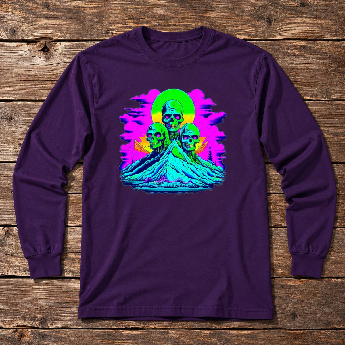 A dark purple long-sleeve t-shirt with a vibrant, colorful graphic print of three skulls on a mountain, displayed flat on a rustic wooden plank background.