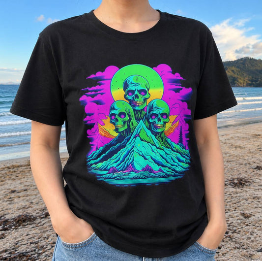 A person wearing a black short-sleeve t-shirt with a vibrant, colorful graphic print of three skulls on a mountain peak under a green and yellow sun, set against a beach background.