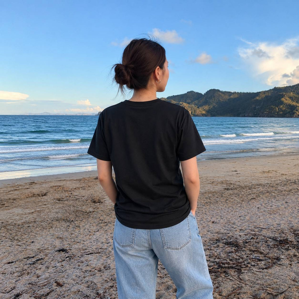 A woman stands on a sandy beach facing the ocean, wearing a plain black short-sleeve t-shirt and light blue jeans.